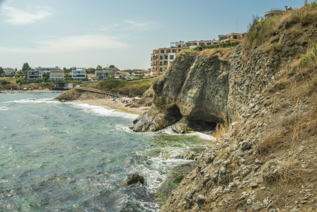 The photo presents waves breaking against rocky coastal cliffs, with a small beach and buildings in the background.