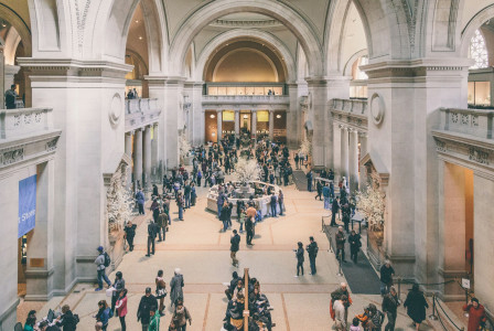 The photo presents a busy museum hall with ornate arches, columns, balconies and visitors gathered around exhibits and information desks.