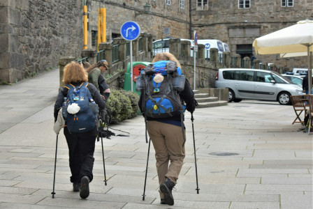 The photo presents two hikers with backpacks and walking poles walking along a stone-paved street.