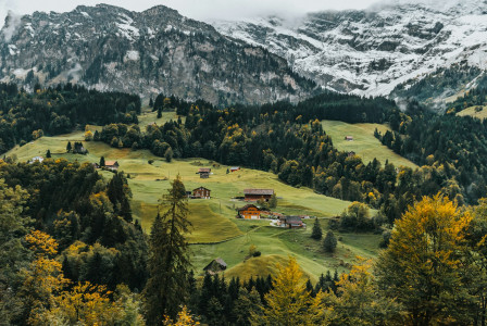 The photo presents scattered wooden houses on green hills, surrounded by forests and autumn foliage beneath snow-covered mountains.