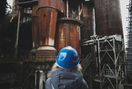 The photo presents a person wearing a blue hard hat facing large rusted industrial structures, metal walkways and staircases.