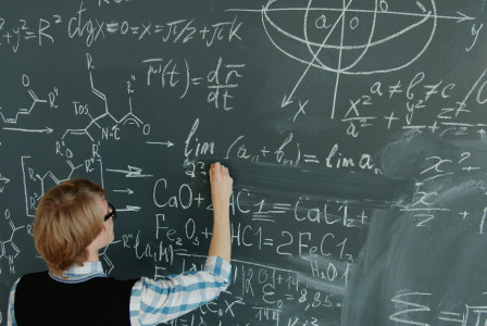 The photo presents a person writing mathematical and chemical equations on a large chalkboard.