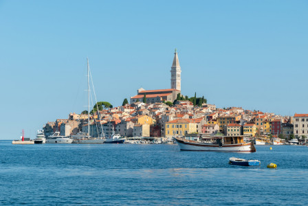 The photo presents boats in a harbour beside a coastal town with colourful buildings and a hilltop church.