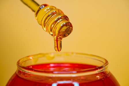 The photo presents honey dripping from a honey dipper into a glass jar.
