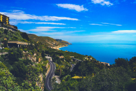 The picture shows a scenic Italian coastline with winding roads, lush green hills, hillside villages, and the blue Mediterranean Sea.