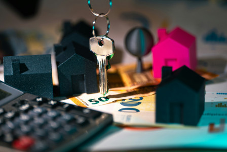 The picture shows house-shaped models, a set of keys, euro banknotes, and a calculator, representing real estate, home ownership, and property financing.