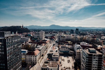 The picture shows an aerial view of an urban landscape in Slovenia, with a mix of modern and historic buildings and mountains in the background.