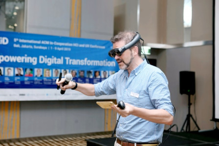 An illustrative photo of  a man in a plaid shirt holds a black and brown corded microphone and wear VR glasses