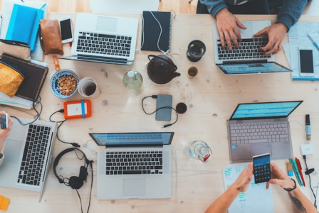 An illustrative photo of a group of people sitting around a table with assorted laptop computers, engaged in a collaborative work session