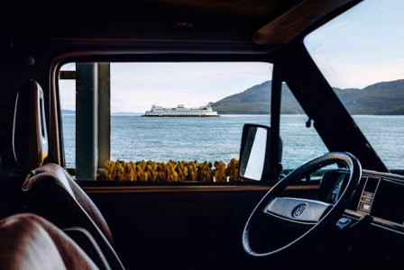 Photo by Pexels An illustrative photo of the interior of a vehicle with a steering wheel in the foreground and a body of water with a ferry in the distance.