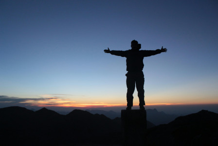 An illustrative photo of a male climber standing on mountain top with outstretched arms at sundown