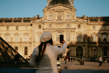 An illustrative photo of a woman in a beret hat taking a picture of the Louvre Museum