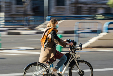 An illustrative photo of a woman in brown jacket wearing a mask and riding on bicycle on road 