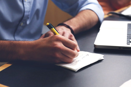 An illustrative photo of a man writing notes on paper with laptop in the background