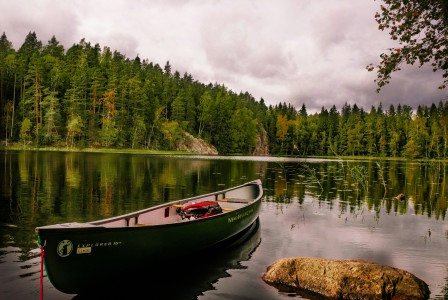 An illustrative photo of a red and white canoe on a lake near green trees under white clouds during the daytime