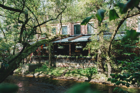 An illustrative photo of a brown wooden house near green trees and river