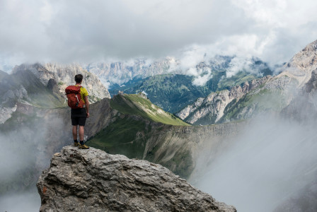 An illustrative photo of a man with red backpack standing on cliff facing mountains.