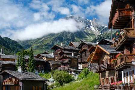 An illustrative photo of brown wooden houses near green trees and mountains.