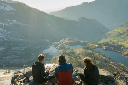 An illustrative photo of three people sitting on cliff overlooking lake and mountains during daytime.