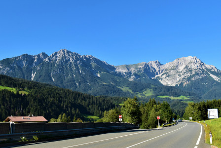 An illustrative photo of a road near green trees and mountains under blue sky.