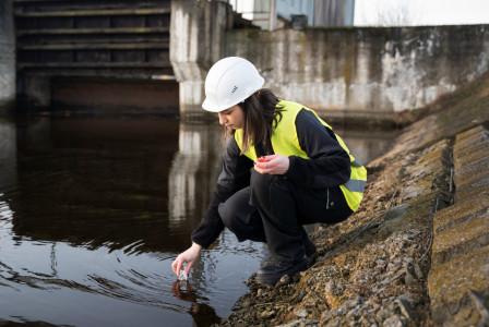 Environmental inspector wearing a safety helmet and high-visibility vest collecting a water sample from a river near a concrete structure.