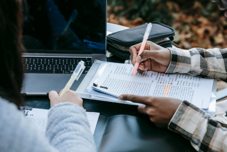 Two people reviewing and highlighting printed documents next to a laptop, collaborating on written materials.