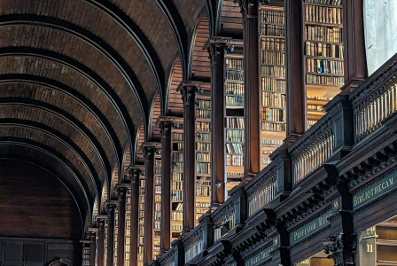 The image shows a grand library interior, with tall wooden bookshelves stretching into the distance and high arched ceilings. Rows of books are neatly arranged along both sides of a long hall. The architecture and layout suggest a historic European library, possibly in an academic setting.