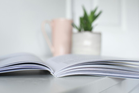 Open book on a table with a mug and small plant in the background.
