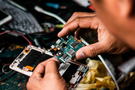 Person repairing a device, holding its internal circuit board over an open device on a cluttered desk