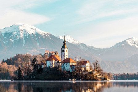 brown-and-white-concrete-building-near-body-of-water-and-mountain-under-white-clouds-and-blue