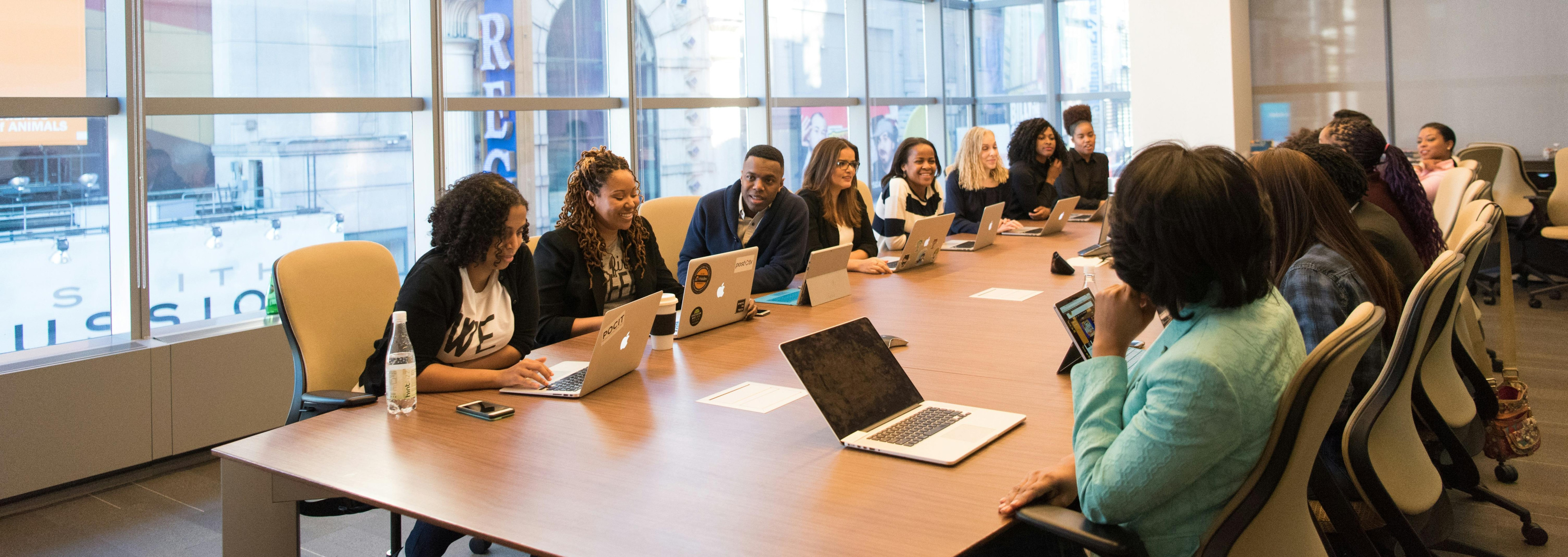 A photo presenting a group of people seated around a large conference table, working on laptops during a meeting.