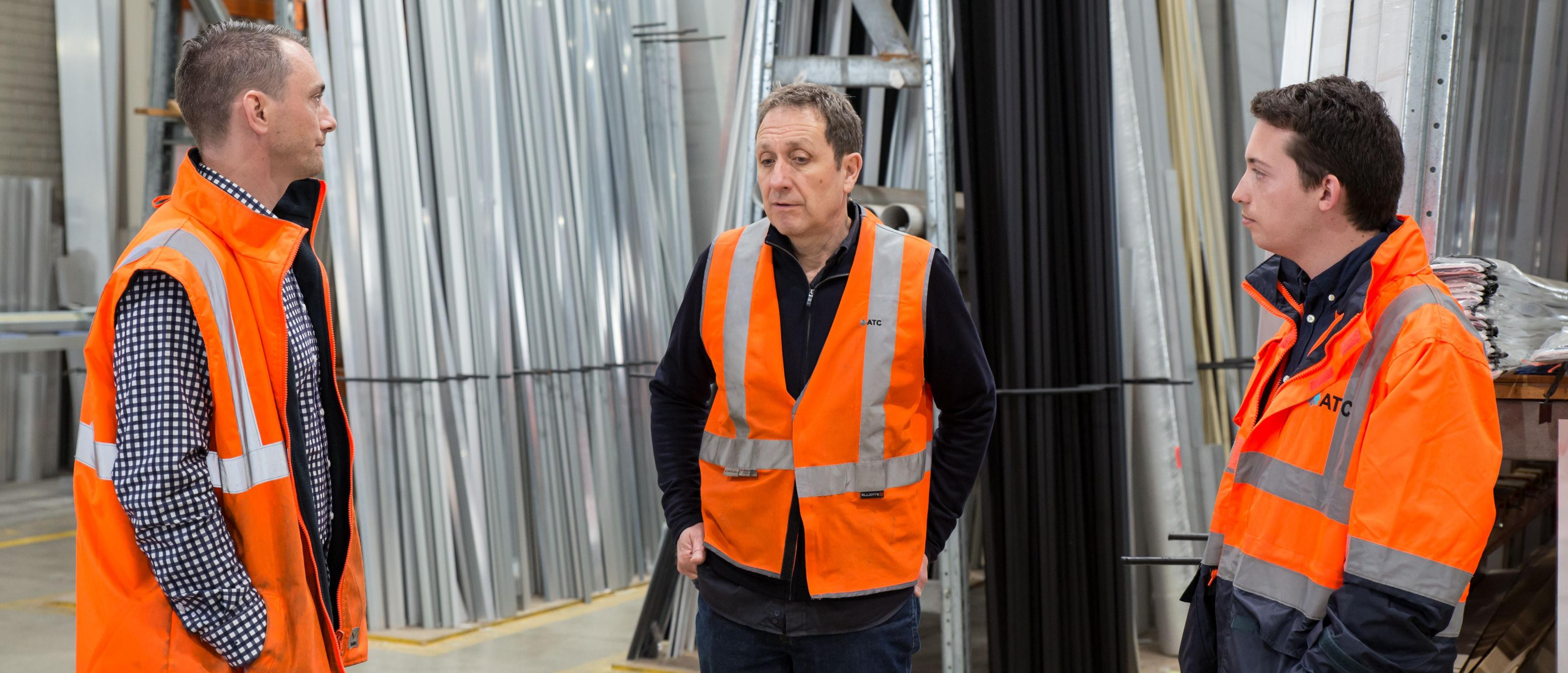 A photo presenting three men in orange safety vests talking in a metal supplies warehouse.