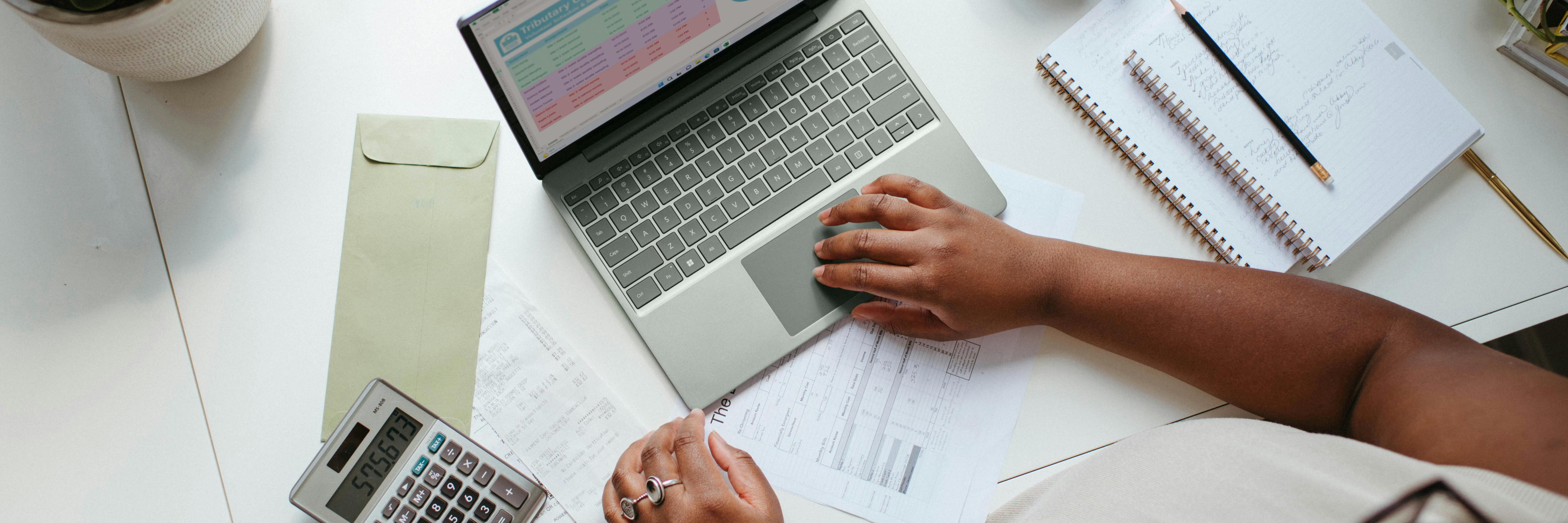 Photo presenting a person working at a desk with a laptop, calculator, documents, and notebook displaying charts and financial data.