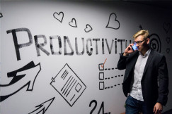 Colour photograph of man walking in front of a white wall with the word "productivity" on it while speaking on the phone, by Andreas Klassen on unsplash.