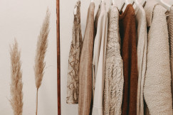 Neutral-toned sweaters, coats, and tops hanging on a clothing rack beside dried pampas grass.