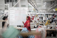 A woman in a red headscarf and a woman in a black dress review notes on a whiteboard in a busy garment production floor.