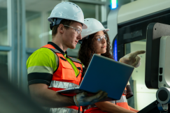 Two workers in safety gear inspect industrial equipment using a laptop.
