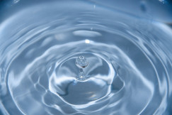 A photo presenting a droplet rebounding from a rippling blue water surface, captured in macro detail.