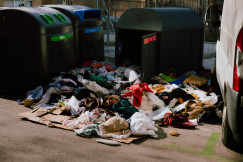 A photo presenting a pile of discarded clothing spilling out beside recycling bins on a street.