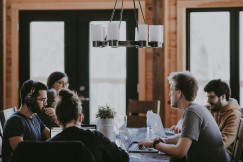 The photo presents a group of people working together with laptops around a table in a collaborative office environment.