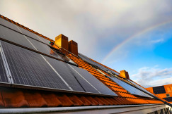 The picture presents solar panels installed on a sloped rooftop beneath a rainbow.