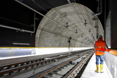 The image shows a railway tunnel equipped with tracks and technical infrastructure, with a construction worker inspecting the underground transport corridor.