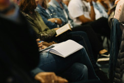 The photo presents people attending a seminar or conference, with one participant taking notes in a notebook.