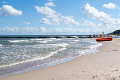 The photo presents waves washing onto a sandy beach, with a red boat and beachgoers under a blue sky with white clouds.