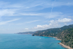 The photo presents a turquoise coastal bay bordered by rugged, forested hills with scattered houses beneath a bright, cloud-filled sky.