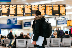 The photo presents a traveller with a backpack and laptop looking at departure boards in a busy terminal.