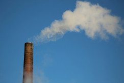 The photo presents a tall brick industrial chimney releasing white smoke into a clear blue sky.