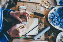 The photo presents a person crafting a small decorative piece at a wooden table covered with tools, moulds and plant cuttings.