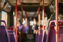 The photo presents the interior of a bus with purple seats, red handrails and seated passengers, illuminated by sunlight.