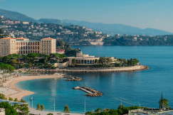The photo presents a coastal resort with hotels, beaches and a marina beside a blue bay, backed by wooded hills and mountains.